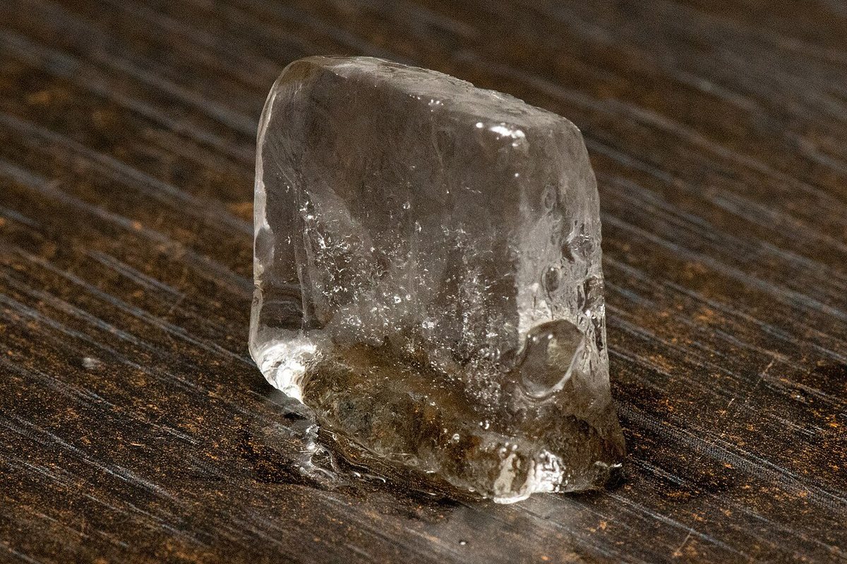 A single ice cube on a wooden table, meltwater pooling around its base